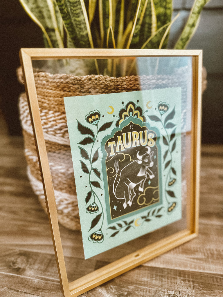 A framed art print in front of luscious house plants in hand woven baskets. A framed art print - the illustration is a depiction of the Taurus zodiac sign. The celestial bull is illustrated floating in a starry night sky, surrounded by whimsical clouds and framed in by folk art flowers. Taurus is hand lettered at the top in a bold, groovy, retro inspired style.
