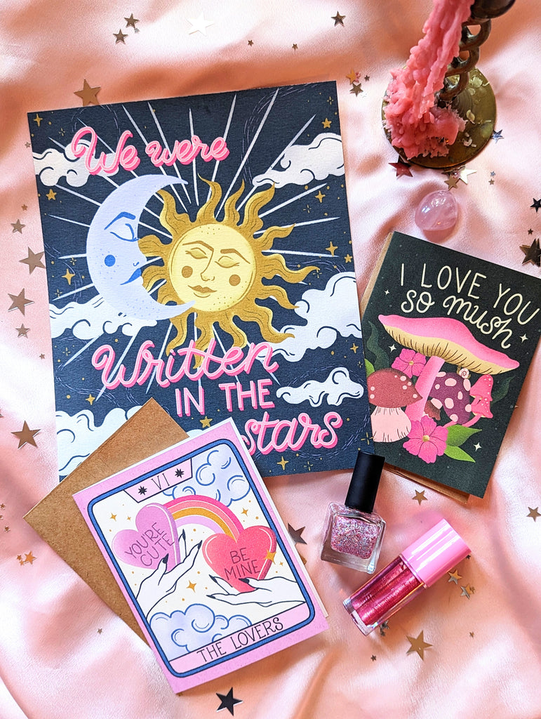Three art prints are shown with scattered star confetti, glitter nail polish, a shining disco ball and crystals all around. The first says “I love you so mush” in hand lettering with mushrooms and fungi all around. The second is reminiscent of The Lovers tarot card, showing hands holding giant candy hearts, with whimsical clouds and twinkles all about. The third says “We Were Written in the stars” in hand lettering script with a beautiful cosmic crescent moon and sun illustration.